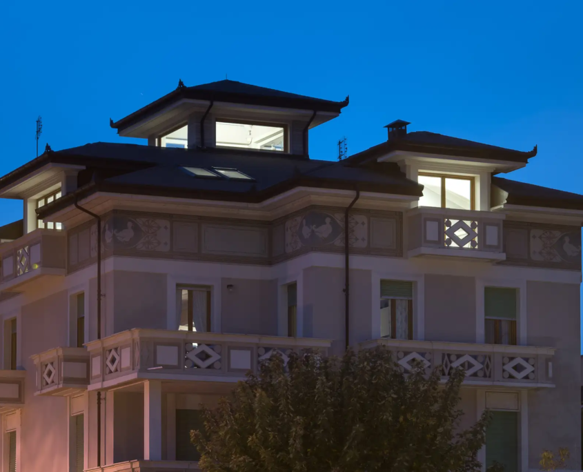 01_cover_progetto_cielo_recuperato vista esterna notturna di palazzo signorile con mansarda illuminata e decorazioni geometriche sui balconi