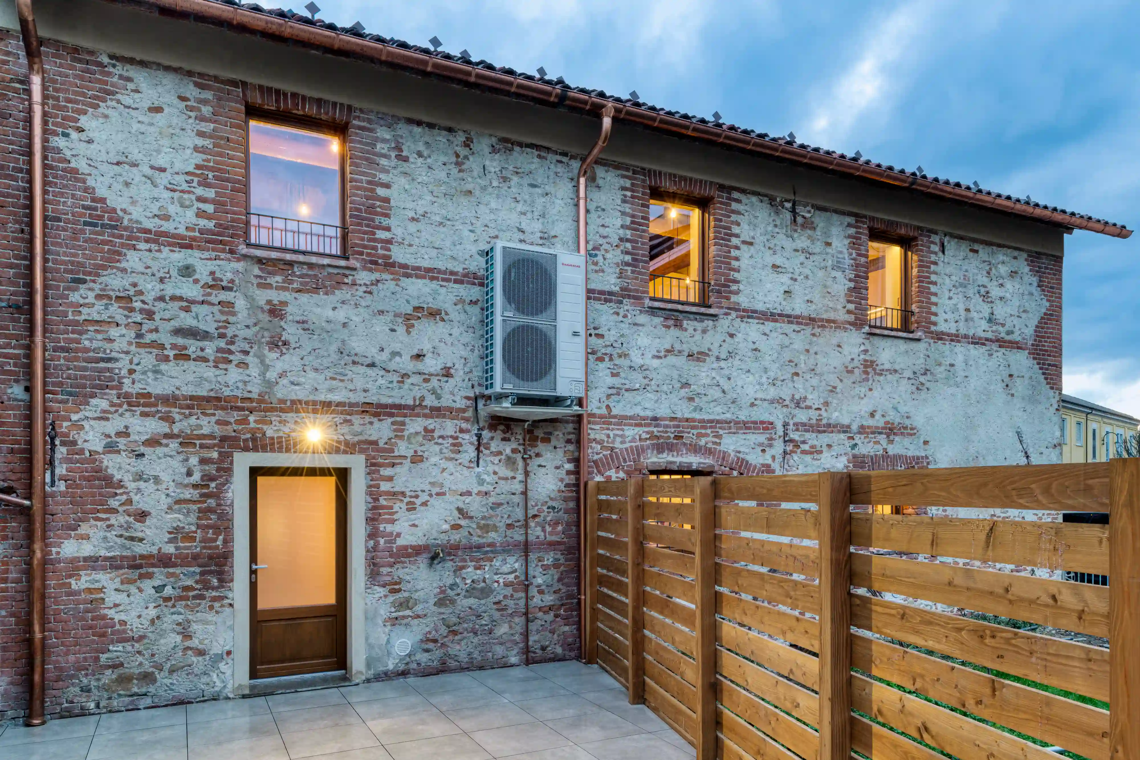 facciata esterna in pietra e mattoni a vista con pompa di calore installata, porta vetrata illuminata e recinzione in legno