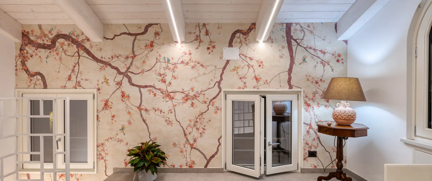 interno con carta da parati a motivo ciliegio in fiore, soffitto con travi in legno chiaro e strisce led integrate, tavolino antico con lampada e pavimento in piastrelle grigie
