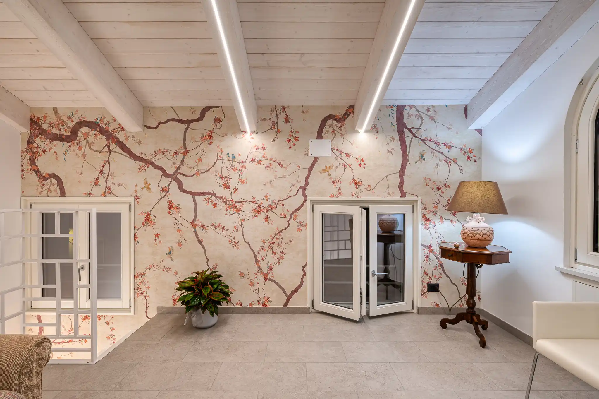 interno con carta da parati a motivo ciliegio in fiore, soffitto con travi in legno chiaro e strisce led integrate, tavolino antico con lampada e pavimento in piastrelle grigie