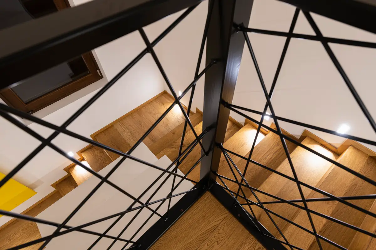 scala interna in legno con ringhiera in metallo nero a trama incrociata, illuminazione led segnapassi e pareti bianche vista dall'alto