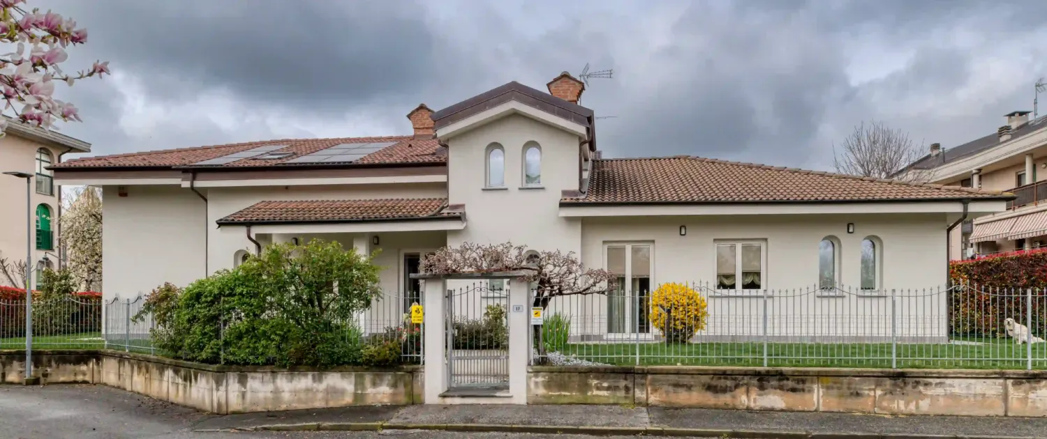 villa singola con facciata bianca, tetto in tegole con pannelli solari, giardino con cespuglio giallo, recinzione metallica e cancello con ramo fiorito in primo piano