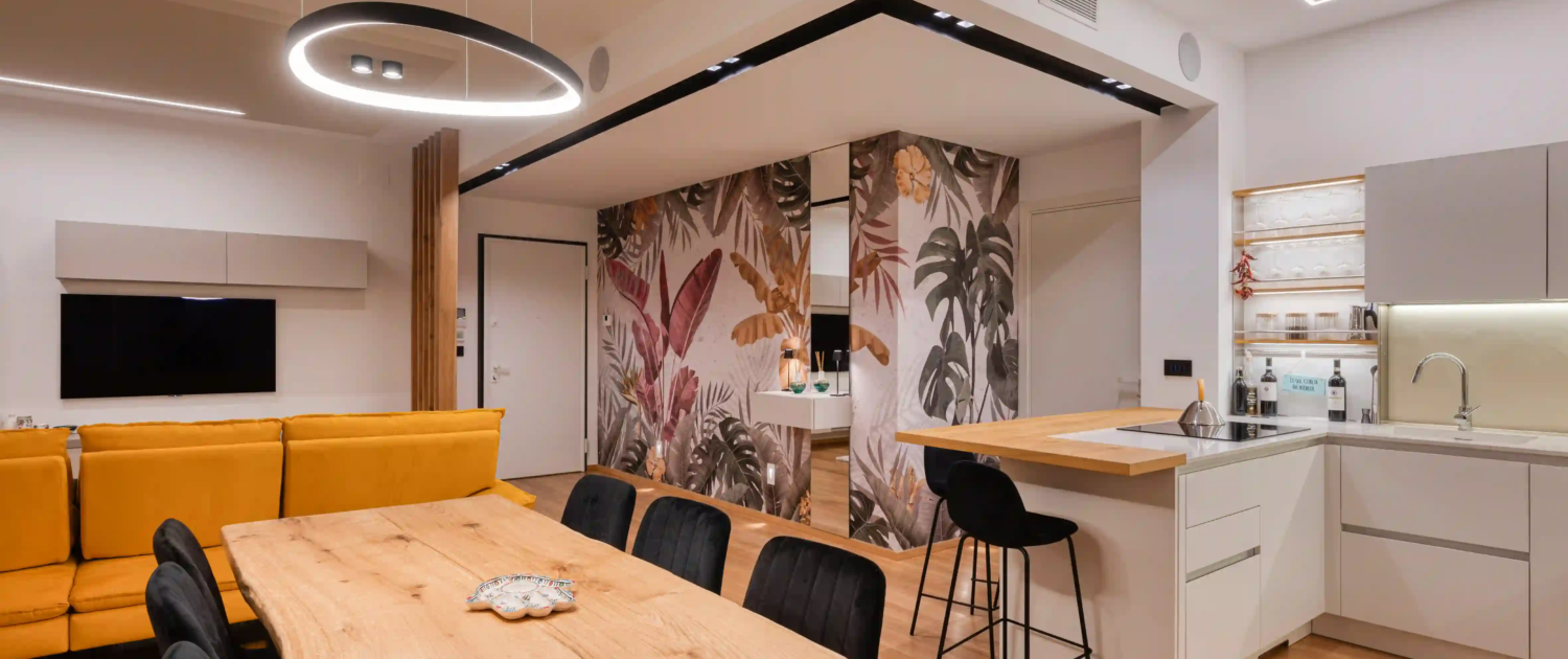 open space con zona living, tavolo in legno e cucina moderna con isola e carta da parati a motivi tropicali