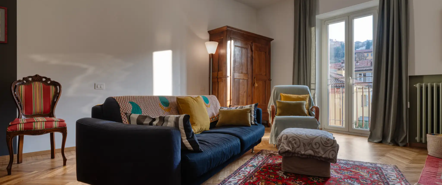 soggiorno con divano blu, tappeto rosso decorato, pavimento in parquet, mobili in legno antico, sedia d epoca e porta finestra con tende scure in edificio residenziale