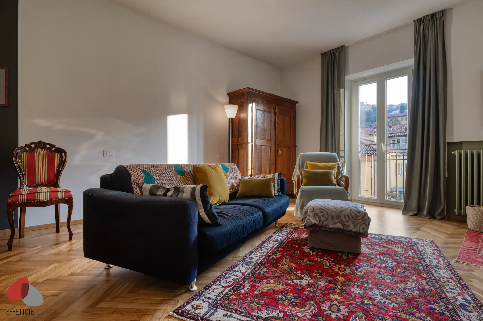 soggiorno con divano blu, tappeto rosso decorato, pavimento in parquet, mobili in legno antico, sedia d epoca e porta finestra con tende scure in edificio residenziale