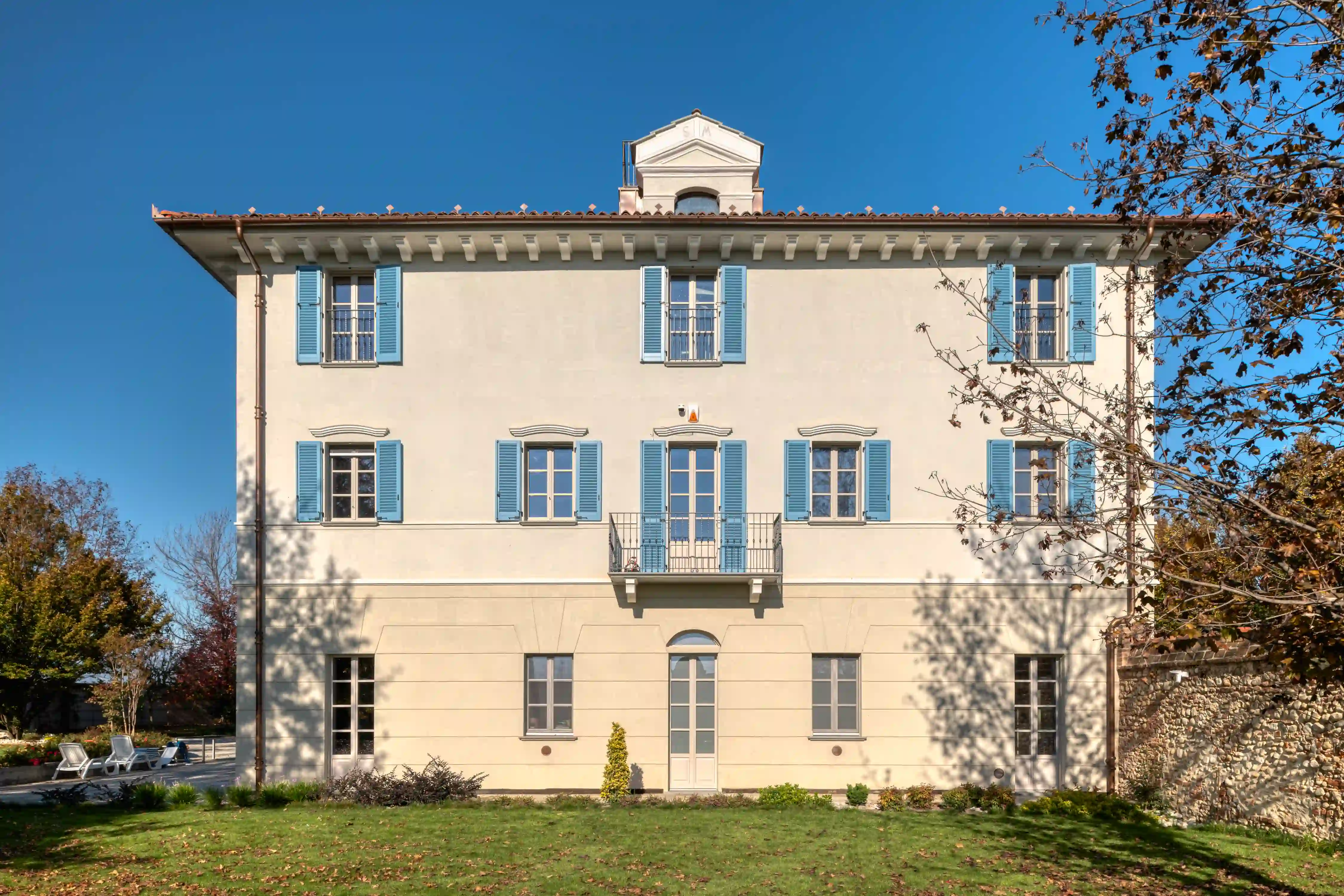 vista frontale del palazzotto restaurato a centallo con facciata chiara, persiane azzurre, giardino curato e cielo azzurro