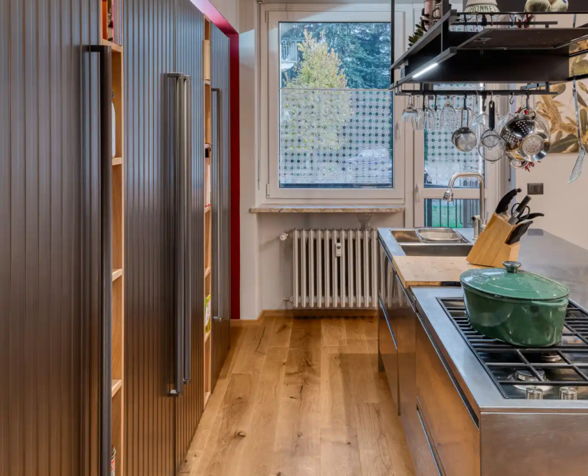 corridoio cucina con armadiature a listelli verticali scuri, isola professionale in acciaio inox e pavimento in parquet
