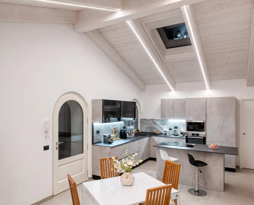 area pranzo con tavolo bianco e sedie in legno, cucina grigia e illuminazione led integrata tra le travi