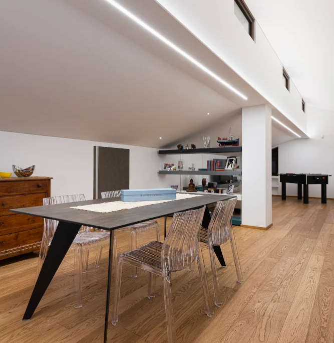 zona pranzo nel sottotetto con tavolo scuro e sedie trasparenti di design con una lunga lama di luce led a soffitto