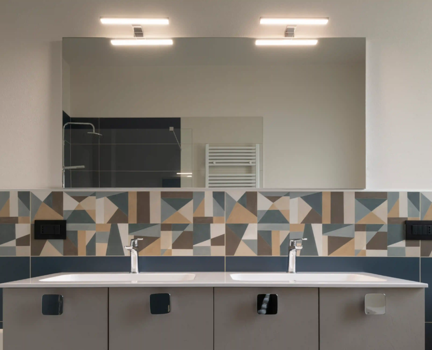 06_immagine_progetto_stella_alpina dettaglio della zona lavabo con due rubinetterie moderne e paraschizzi in piastrelle con pattern geometrico multicolore