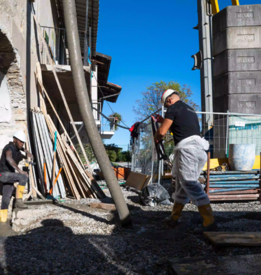 cantiere edile esterno con operai, getto di cemento tramite pompa, gru con contrappesi sullo sfondo, stivali gialli e cielo azzurro