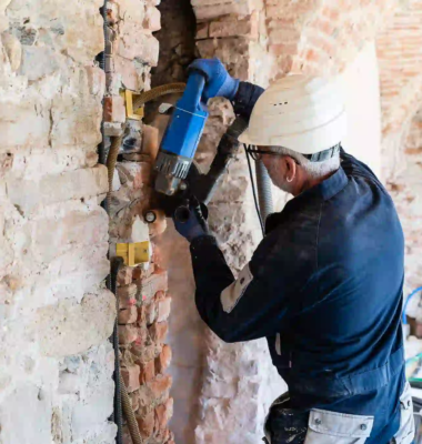 operaio con casco bianco e tuta blu che utilizza un elettroutensile su una parete in mattoni a vista durante lavori di ristrutturazione interna