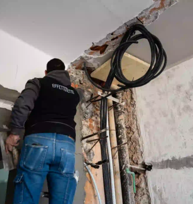 operaio visto di spalle con maglia effettotetto e jeans, impegnato in lavori su una parete con cavi e tubi corrugati a vista durante una ristrutturazione interna