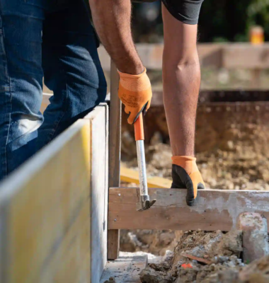 operaio che utilizza un martello su assi di legno in un cantiere edile esterno, guanti arancioni e terreno in primo piano