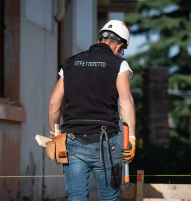 operaio visto di spalle con maglia effettotetto, casco bianco, cintura portautensili e martello in un cantiere edile esterno