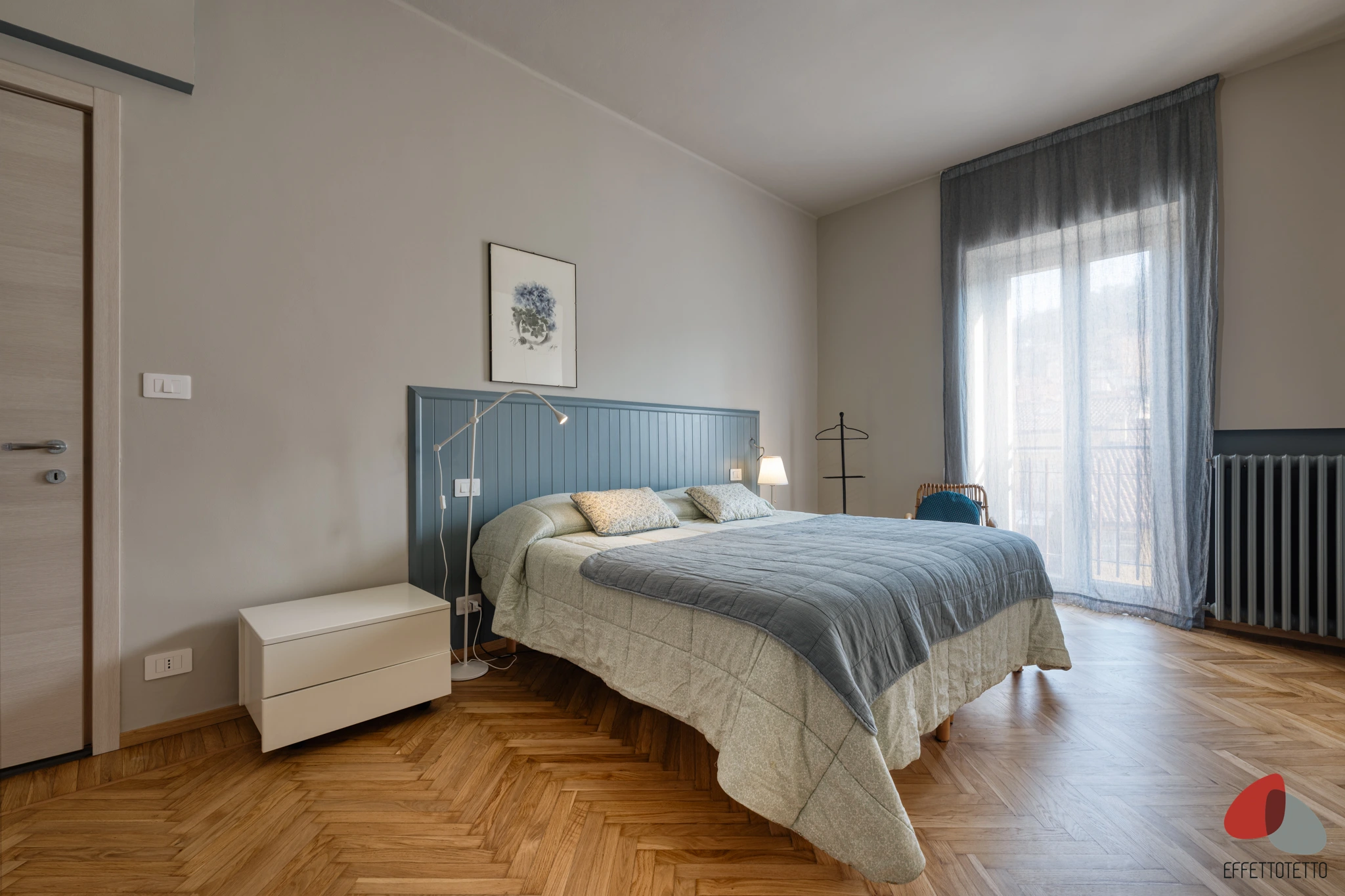 camera da letto luminosa con parquet, testiera azzurra e ampia finestra con tende leggere